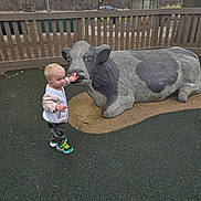 Odin joined the competition — help win amazing prizes! boy, camouflage_pants, car_in_background, child, cow_statue, fence, ground_cover, hand, jacket, outdoor, overcast_sky, park, playground, portrait, rubber_play_surface, sculpture, sneakers, statue, toddler, trees