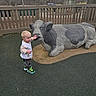boy, camouflage_pants, car_in_background, child, cow_statue, fence, ground_cover, hand, jacket, outdoor, overcast_sky, park, playground, portrait, rubber_play_surface, sculpture, sneakers, statue, toddler, trees
