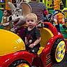 Odin is registered to the contest to win money with this photo: arcade, blue_wheel, bright_colors, carousel_horse, cartoon_mouse, child, colorful, family, indoor_playground, kiddie_ride, neutral_expression, parent, plastic_toy, play_area, red_car, striped_shirt, toddler, woman, wristband, yellow_seat