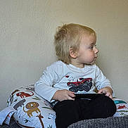 Odin is registered to the contest to win money with this photo: animal_pillow, black_pants, blonde_hair, candid, casual_clothing, child, curly_hair, home, indoor, looking_away, monster_truck_graphic, neutral_background, plush_pillow, seated, side_profile, small_child, toddler, toy, white_shirt, young_child