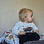 Odin is registered to the contest to win money with this photo: animal_pillow, black_pants, blonde_hair, candid, casual_clothing, child, curly_hair, home, indoor, looking_away, monster_truck_graphic, neutral_background, plush_pillow, seated, side_profile, small_child, toddler, toy, white_shirt, young_child