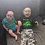 two_children, baby, toddler, sibling, smiling, laughing, couch, living_room, onesie, pants, barefoot, car_prints, graphic_shirt, remote_control, carpet, portrait, happy, infant, cute, playful