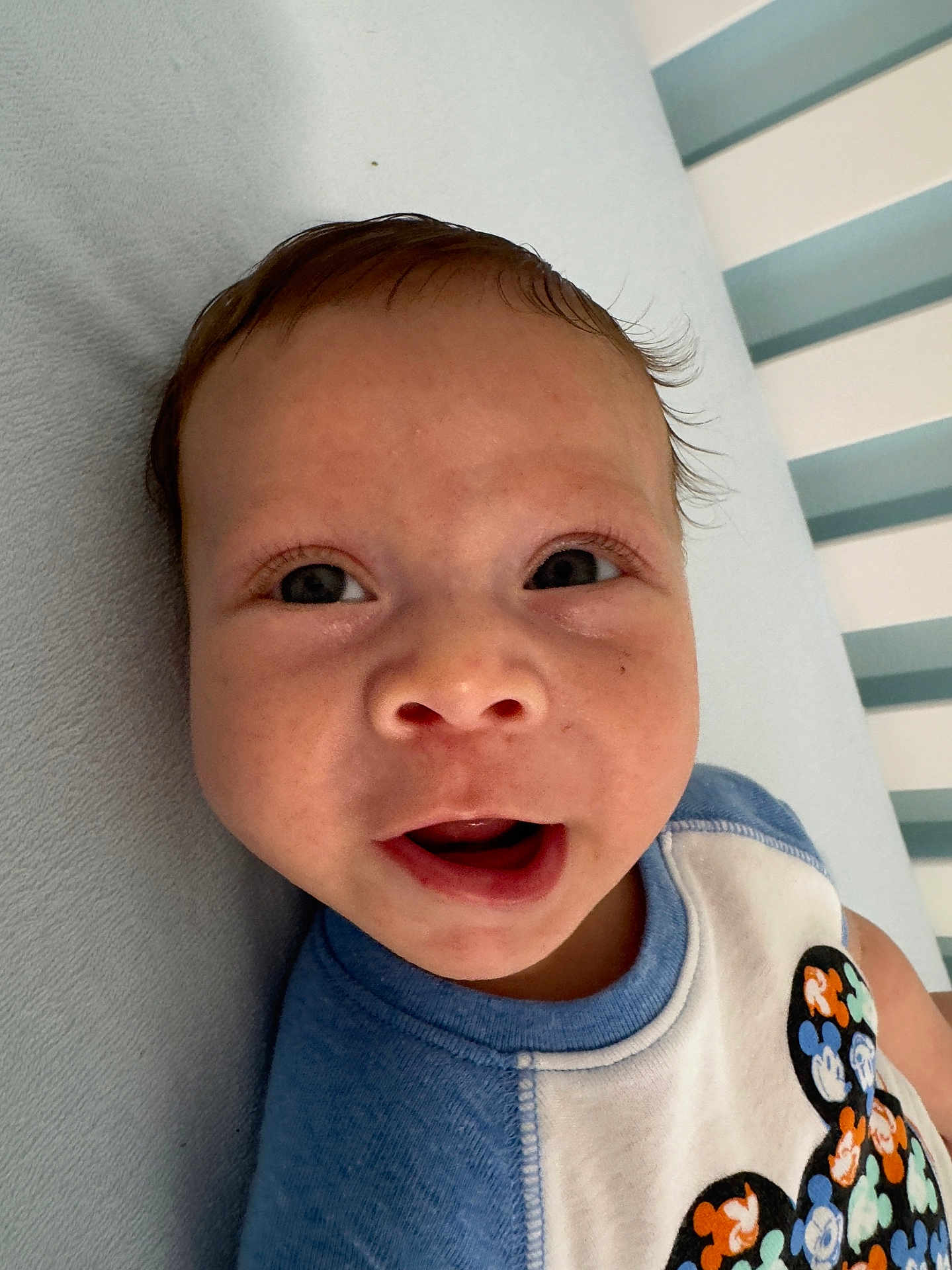 Ezekiel is registered to the contest to win money with this photo: baby, child, face, smiling, blue_clothing, mickey_mouse, pattern, striped_background, infant, cute, portrait, happy, lying_down, soft_surface, short_hair, indoors, close_up, person, toddler, apparel
