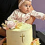 baby, infant, cake, floral_decor, headband, pink_clothing, arm, holding, celebration, icing, cross_symbol, living_room, couch, cake_plate, hands, eyes, portrait, dress, remote_control, flower