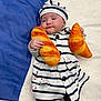 baby, infant, striped_clothing, hat, plush_toy, bread_toy, croissant_toy, soft_surface, blue_background, white_background, cute, child, lying_down, adorable, smiling, indoor, person, cozy, fashion, portrait