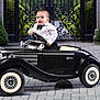 Liam is registered to the contest to win money with this photo: baby, black_car, bushes, child, cute, formal_clothing, gate, greenery, iron_gate, miniature_car, outdoor, pavement, person, portrait, smiling, steering_wheel, toy_car, tuxedo, vintage_car, white_walls