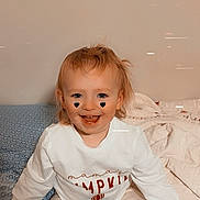 Aniya is registered to the contest to win money with this photo: toddler, child, smiling, face, heart_shape, cheeks, white_shirt, text_on_clothing, pants, bed, blanket, blue_blanket, white_blanket, indoor, happy, sitting, cute, young_child, freckles, person