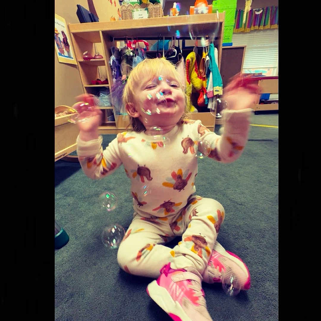 Aniya is registered to the contest to win money with this photo: blond_hair, bubbles, carpet, child, clothing, colorful, furniture, happy, indoor, motion_blur, pajamas, person, pink_sneakers, playful, playroom, sitting, smiling, toddler, toys, young_child