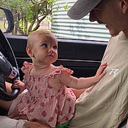 Avery is registered to the contest to win money with this photo: baby, child, man, car_interior, steering_wheel, watch, cap, pink_dress, window, tree, seat, hand, smile, face, interaction, curiosity, affection, casual_clothing, daylight, portrait