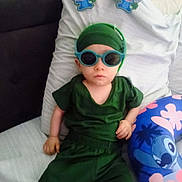 Xavier is registered to the contest to win money with this photo: child, baby, infant, sunglasses, green_outfit, headwrap, pillow, bed, couch, sticker, stitch_character, cartoon, blue_pillow, floral_pattern, portrait, indoor, cute, serious_expression, relaxed, seated