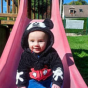 Mélio participe au concours pour gagner de l'argent avec cette photo : baby, child, slide, playground, hoodie, mickey_mouse, outdoor, smiling, sunny, grass, blue_sky, house, fence, clothing, person, cute, sitting, fun, daytime, park