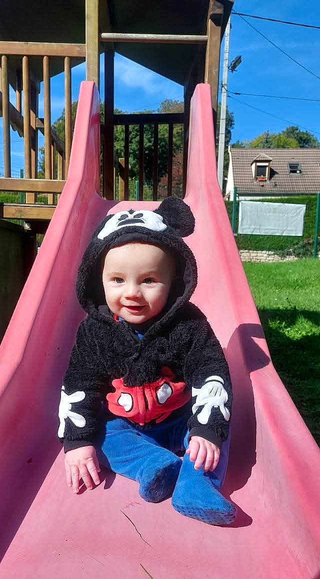 Mélio participe au concours pour gagner de l'argent avec cette photo : baby, child, slide, playground, hoodie, mickey_mouse, outdoor, smiling, sunny, grass, blue_sky, house, fence, clothing, person, cute, sitting, fun, daytime, park