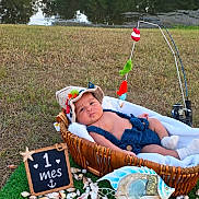 Angel is registered to the contest to win money with this photo: baby, infant, child, portrait, outdoor, grass, pond, basket, bassinet, sunhat, knitwear, blue_clothing, mobile_toy, fishing_theme, toy_fish, seashells, chalkboard, one_month, lying_down, baby_photography
