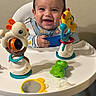 baby, high_chair, smiling, toys, infant, playtime, happy, child, face, clothing, indoor, mirror, pacifier, colorful, plastic_toys, seat, cute, young_child, background_wall, playful