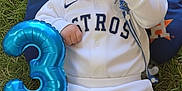 Derek Caleth is registered to the contest to win money with this photo: baby, smiling, grass, balloon, number_three, baseball_outfit, cap, white_clothing, orange_pillow, blue_pillow, houston_astros, pacifier, celebration, infant, happy, outdoor, sportswear, cute, lying_down, child