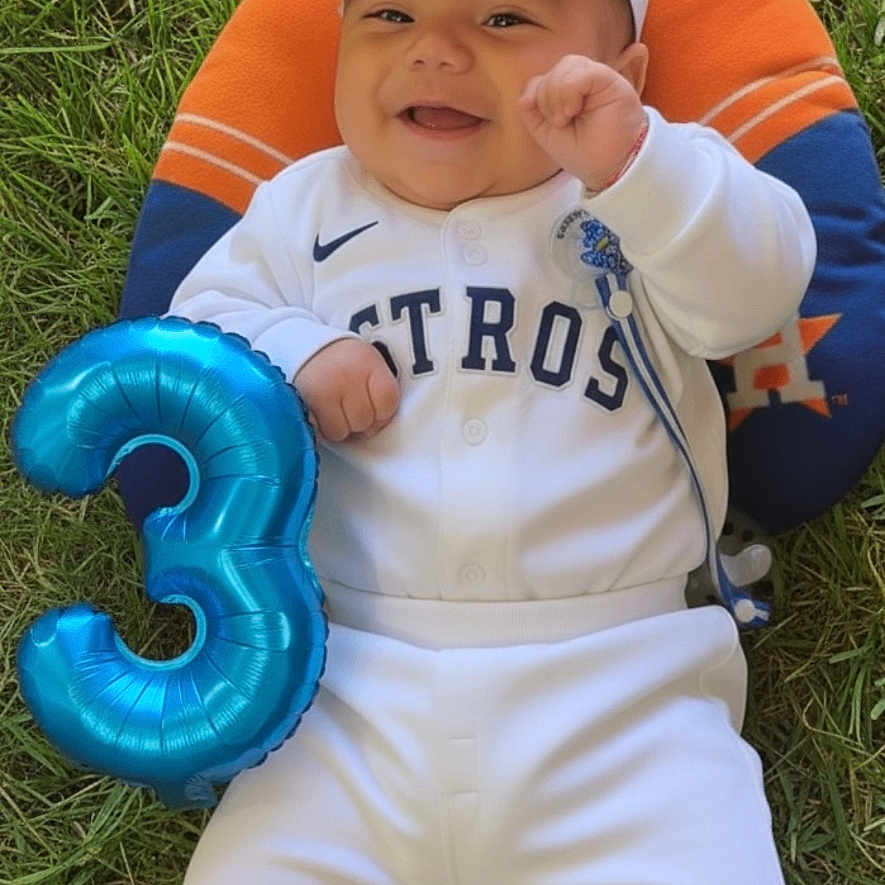 Derek Caleth is registered to the contest to win money with this photo: baby, balloon, baseball_outfit, blue_pillow, cap, celebration, child, cute, grass, happy, houston_astros, infant, lying_down, number_three, orange_pillow, outdoor, pacifier, smiling, sportswear, white_clothing