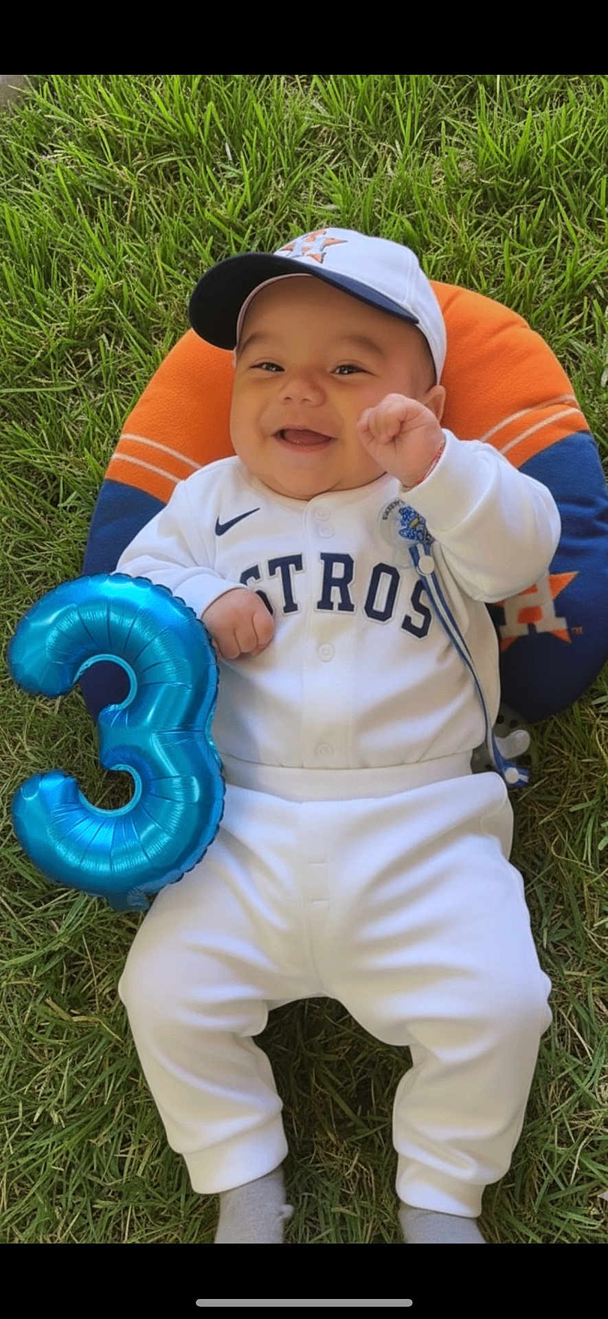 Derek Caleth is registered to the contest to win money with this photo: baby, smiling, grass, balloon, number_three, baseball_outfit, cap, white_clothing, orange_pillow, blue_pillow, houston_astros, pacifier, celebration, infant, happy, outdoor, sportswear, cute, lying_down, child