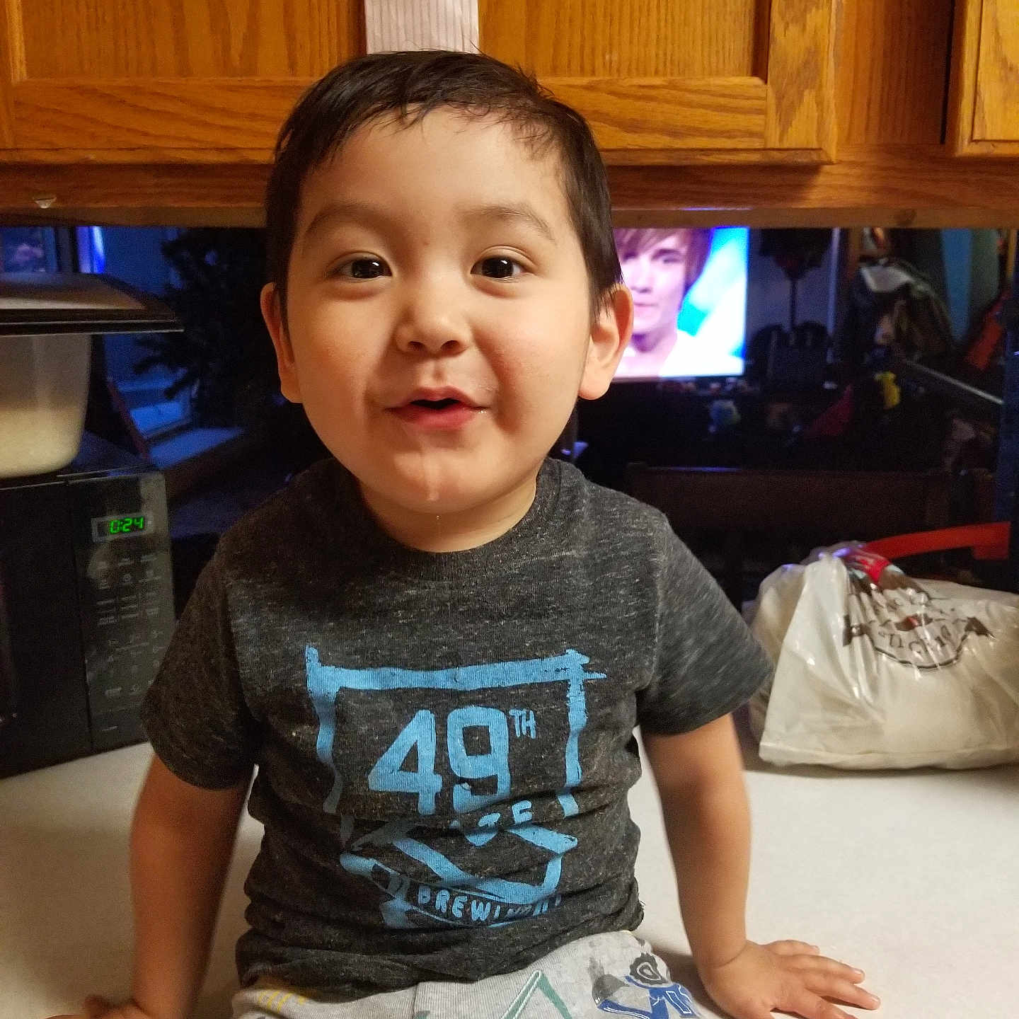 Kohen Combs joined the competition — help win amazing prizes! appliance, boy, casual_clothing, child, counter, drool, expression, face, furniture, home, indoor, kitchen, microwave, pants, playful, sitting, smiling, television, tshirt, wooden_cabinets