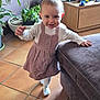 child, toddler, baby, smiling, dress, socks, standing, living_room, plants, potted_plant, toys, wooden_drawer, couch, floor_tiles, happy, cute, hand, face, eyes, portrait