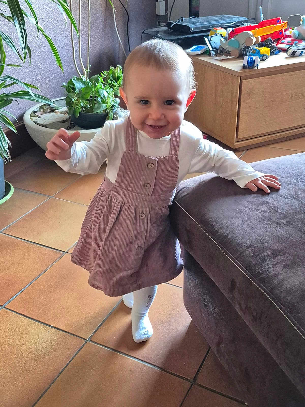Kalya a rejoint le concours — aidez-le/la à gagner de superbes lots ! child, toddler, baby, smiling, dress, socks, standing, living_room, plants, potted_plant, toys, wooden_drawer, couch, floor_tiles, happy, cute, hand, face, eyes, portrait