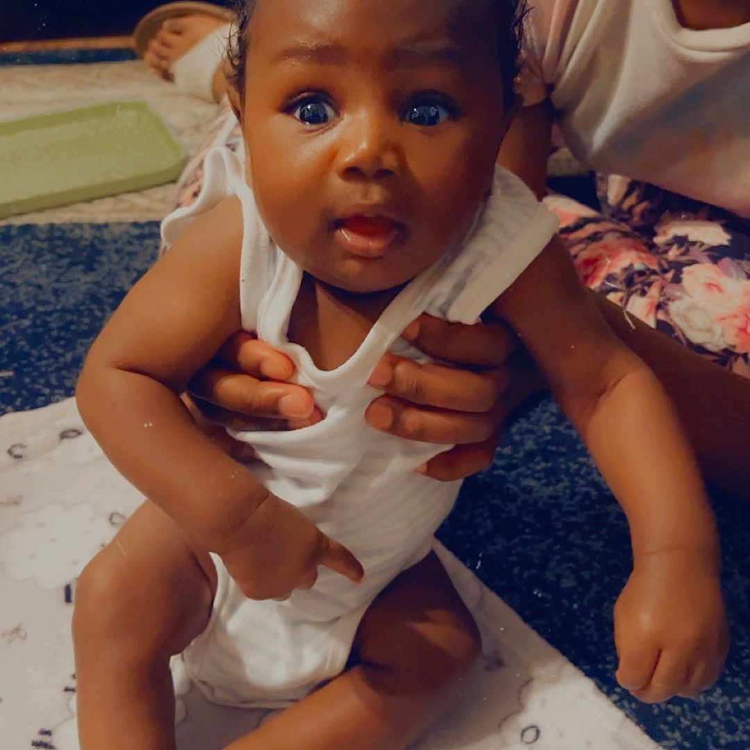 Khyzun Cooper is registered to the contest to win money with this photo: baby, blanket, carpet, child, curly_hair, cute, expression, face, floor, foot, hands, holding, indoor, infant, pattern, person, sitting, skin, toe, white_clothing