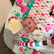 Amelia is registered to the contest to win money with this photo: baby, infant, smiling, play_mat, toy, colorful, pink, teal, floral_pattern, clothing, cute, child, happy, lying_down, indoor, soft_texture, four_months, playtime, person, face