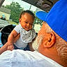 baby, child, man, adult, car_interior, steering_wheel, blue_cap, white_shirt, bib, smile, happy, facial_expression, holding, hand, window, trees, outdoor, vehicle, person, interaction