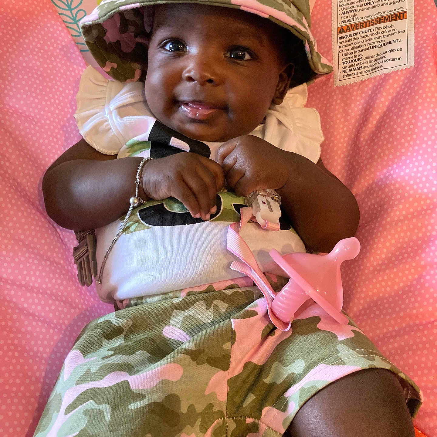 Ka Laeyah is registered to the contest to win money with this photo: accessory, baby, bracelet, camo_clothing, child, closeup, cushion, cute, face, green, happy, hat, indoors, infant, pacifier, person, pink, polka_dot, seated, smiling