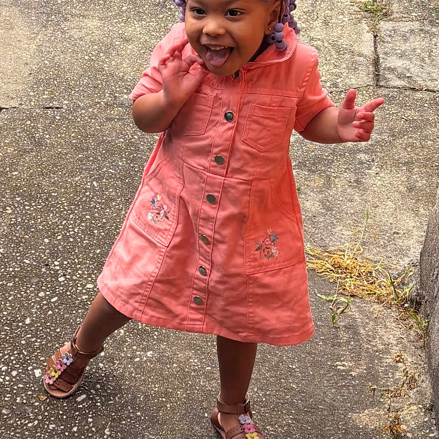 Tania is registered to the contest to win money with this photo: toddler, child, girl, smiling, playful, outdoor, sidewalk, concrete, grass, dress, coral, sandals, braids, purple_beads, happy, person, standing, cute, young, joyful