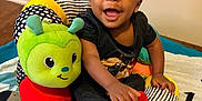 Markel is registered to the contest to win money with this photo: baby, child, smiling, indoor, plush_toy, caterpillar, colorful, sitting, red_socks, black_clothing, playful, happy, face, person, floor, door, carpet, toy, infant, cute