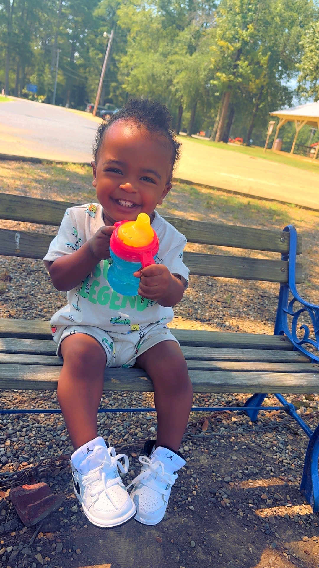 Markel joined the competition — help win amazing prizes! toddler, child, bench, sippy_cup, smile, outdoor, park, shoes, white_sneakers, clothing, happy, person, daylight, trees, gravel, playground, nature, baby, cute, fun
