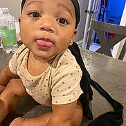 Lee is registered to the contest to win money with this photo: baby, child, tongue_out, polka_dot_clothing, head_wrap, table, indoor, wooden_table, chair, water_bottle, kitchen, playful, cute, person, skin, face, eyes, earring, sitting, infant