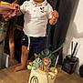 birthday_cake, cake, candle, celebration, child, curtains, decorations, gold_balls, hand, indoor, lion_figurine, monkey_figurine, party, person, plants, shorts, smile, table, toddler, white_shirt