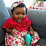 baby, child, smiling, dress, headband, toy, sitting, couch, indoor, happy, curly_hair, barefoot, floral_pattern, person, infant, cute, colorful, playful, young, portrait