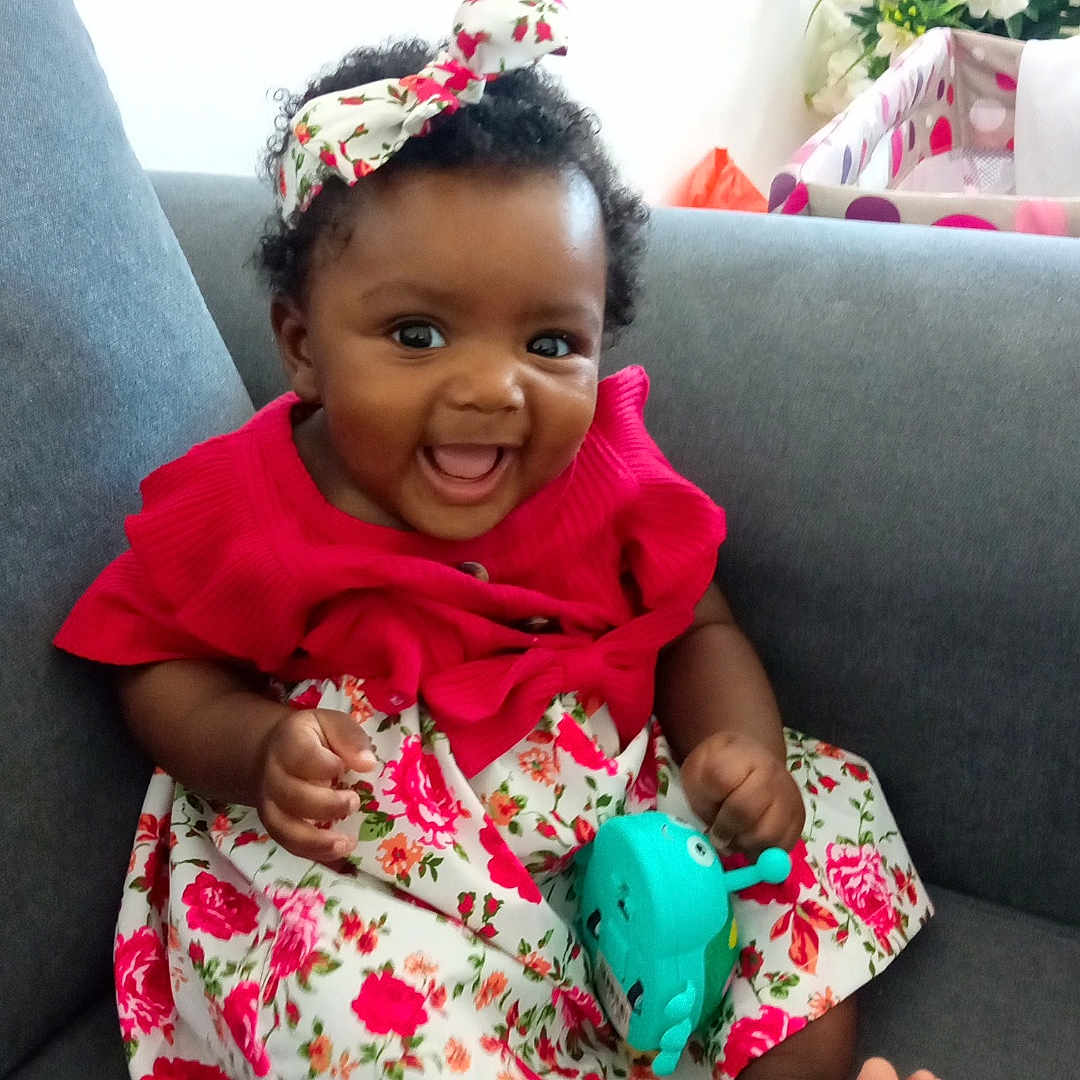 Soriah participe au concours pour gagner de l'argent avec cette photo : baby, barefoot, child, colorful, couch, curly_hair, cute, dress, floral_pattern, happy, headband, indoor, infant, person, playful, portrait, sitting, smiling, toy, young