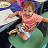 child, toddler, smiling, pigtails, pink_shirt, coloring, crayon, paper, chair, green_chair, table, shoes, sneakers, indoor, classroom, happy, person, cute, drawing, playful