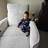 Azariah is registered to the contest to win money with this photo: baby, child, armchair, furniture, indoor, person, clothing, onesie, patterned_clothing, socks, carpet, wall, power_outlet, cabinet, curious, relaxed, seated, home, cozy, portrait