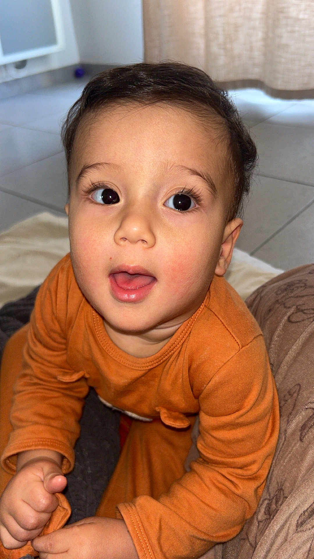 Nahyl a rejoint le concours — aidez-le/la à gagner de superbes lots ! baby, blanket, child, curly_hair, cute, expression, face, floor, hand, happy, home, indoor, long_sleeve, orange_clothing, person, portrait, sitting, skin, toddler, young