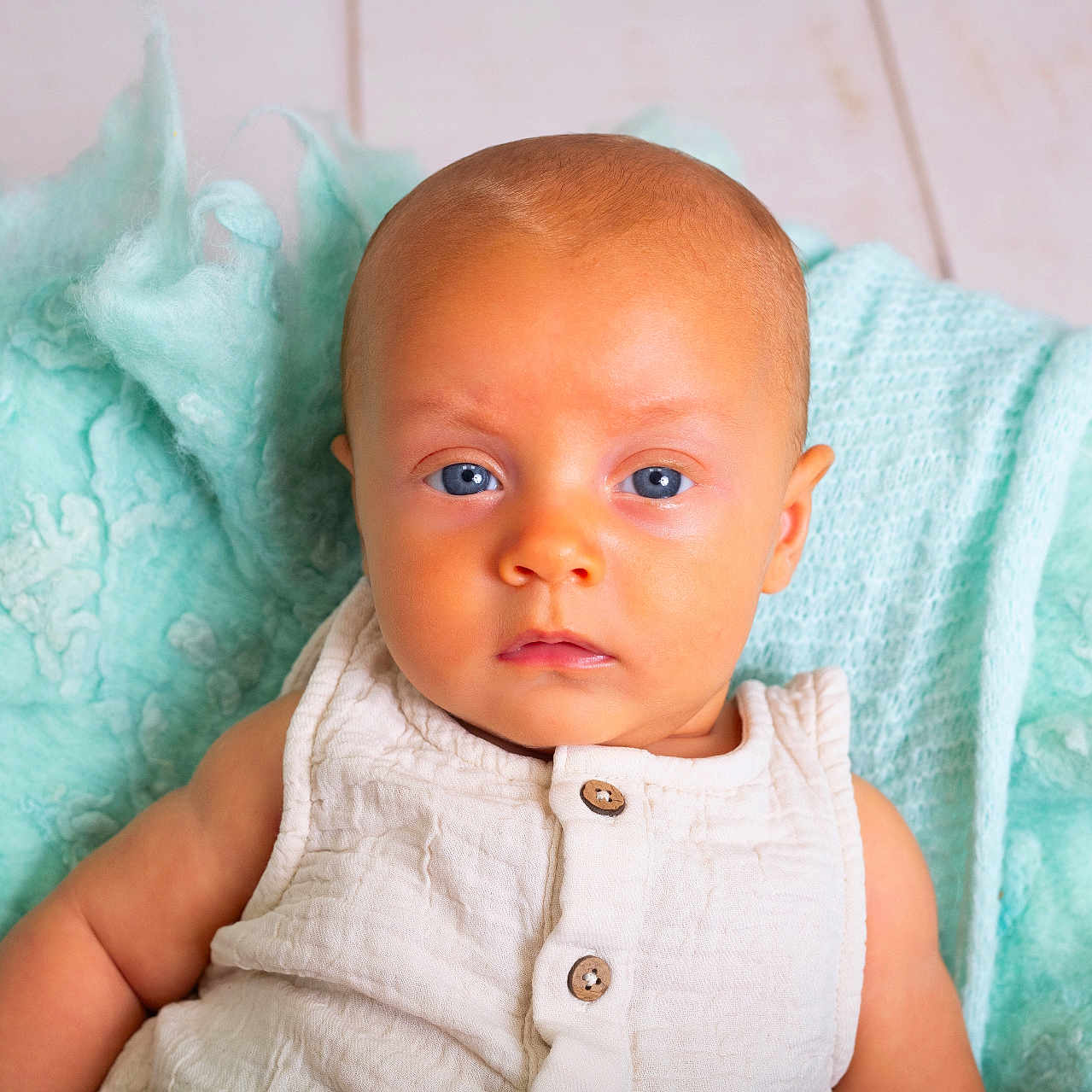 Léonis a rejoint le concours — aidez-le/la à gagner de superbes lots ! baby, blanket, blue_eyes, calm, closeup, cute, face, floor, head, indoor, infant, newborn, person, portrait, sitting, skin, soft_texture, white_clothing, wooden_floor, young_child