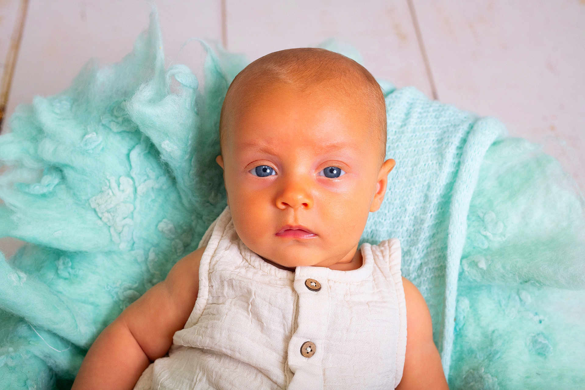 Léonis a rejoint le concours — aidez-le/la à gagner de superbes lots ! baby, infant, blue_eyes, white_clothing, blanket, soft_texture, portrait, face, head, skin, cute, young_child, person, indoor, floor, wooden_floor, calm, sitting, closeup, newborn