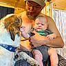 Elvis Presley is registered to the contest to win money with this photo: baby, dog, man, glasses, cap, indoor, wooden_wall, plaid_pants, pet, infant, shirtless, paw, holding, curtain, facial_hair, onesie, collar, home, interaction, cabin