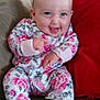baby, smiling, infant, floral_onesie, couch, red_pillow, happy, sitting, indoors, cute, child, patterned_fabric, joyful, face, person, home, cozy, small_hands, footed_pajamas, cheerful