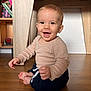 baby, child, smiling, sitting, floor, wooden_floor, long_sleeve_shirt, pants, indoors, home, furniture, shelf, toy, happy, face, person, cute, infant, young_child, natural_light