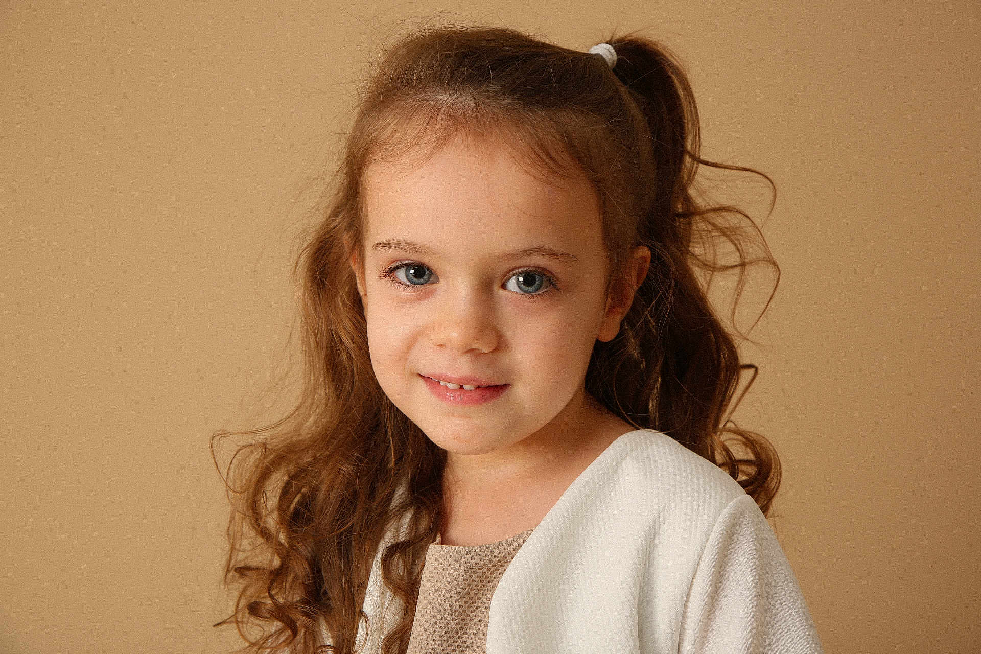 Maëline a rejoint le concours — aidez-le/la à gagner de superbes lots ! child, girl, portrait, smile, curly_hair, pony_tail, white_cardigan, beige_background, face, person, young, cute, innocence, soft_lighting, indoor, fashion, head_and_shoulders, happy, casual_clothing, studio_photo