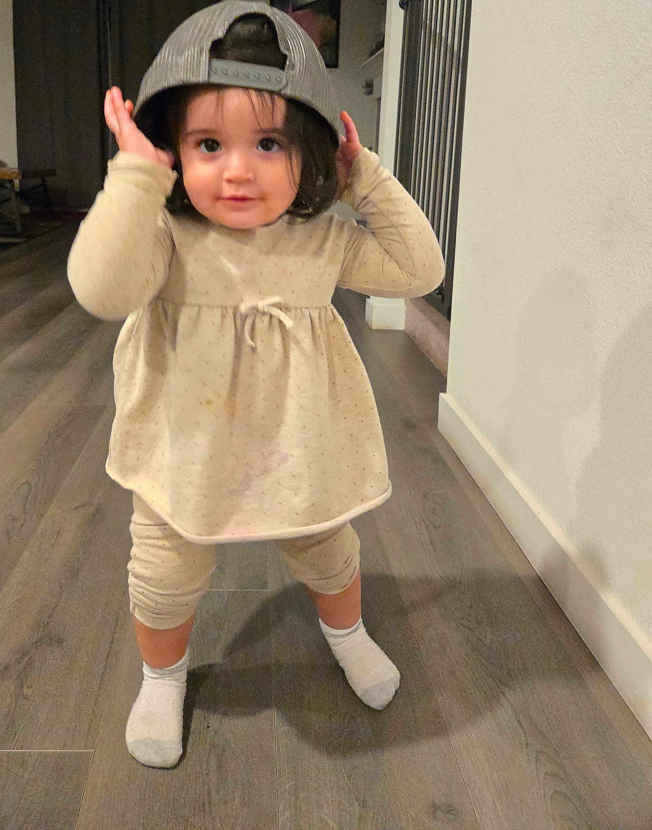 Melissa Arellano joined the competition — help win amazing prizes! baseball_cap, beige_clothing, casual, child, cute, floor, hair, home, indoor, lighting, long_hair, person, playful, smiling, socks, standing, toddler, wall, wood_floor, young_child
