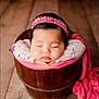 baby, sleeping, headband, wooden_bucket, pink_blanket, floral_pattern, infant, close_up, portrait, cozy, peaceful, hands_under_chin, indoors, flooring, cute, newborn, soft_lighting, resting, child, adorable