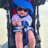 baby, stroller, blue_hat, sunglasses, smile, sunny_day, toy, outdoor, clothing, legs, shoes, seat_belt, child, drool, plastic_toy, black_stroller, fabric, shadow, person, happy
