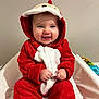 Lylia a rejoint le concours — aidez-le/la à gagner de superbes lots ! adorable, baby, child, clothing, cozy, cushion, cute, face, hands, happy, hood, indoor, infant, playful, portrait, red_onesie, sitting, smile, white_background, young_child