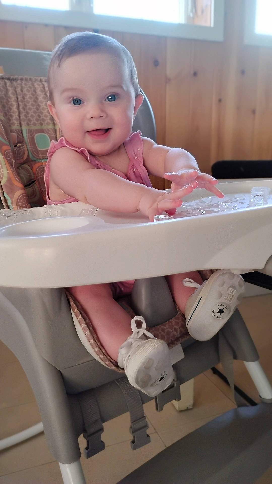 arm, baby, child, expression, face, floor, footwear, hand, happy, high_chair, indoor, infant, person, pink_clothing, seat, shoe, smile, table, window, wood_paneling