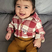 Sara is registered to the contest to win money with this photo: baby, child, smiling, red_shirt, checkered_shirt, mustard_pants, white_socks, couch, gray_couch, happy, indoor, portrait, infant, clothing, sitting, cute, young_child, face, person, casual